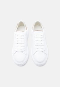 Paire de baskets basses blanches impeccables avec accents de talon en daim beige, lacets blancs et semelles en caoutchouc texturé sur un fond clair.