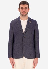 Uomo che indossa un blazer su misura blu scuro sopra una maglietta bianca a girocollo e pantaloni beige chiaro, in piedi davanti a uno sfondo semplice.