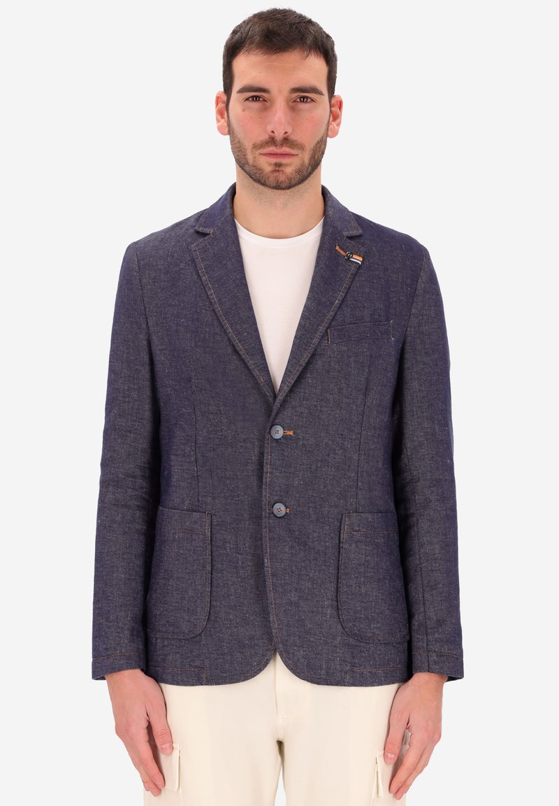 Uomo che indossa un blazer su misura blu scuro sopra una maglietta bianca a girocollo e pantaloni beige chiaro, in piedi davanti a uno sfondo semplice.