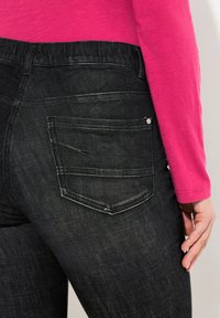 Jeans de mezclilla negra con un diseño ajustado, que cuentan con bolsillos traseros y un ligero desgaste. Combinados con una camiseta de manga larga rosa.