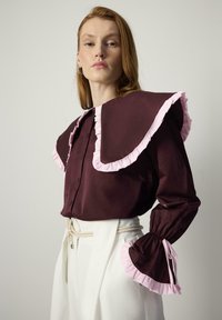 Bordeaux blouse met grote, rimpelige roze kraag en mouwen; knoopsluiting aan de voorkant; gecombineerd met witte hoge taille broeken en een strikriem.