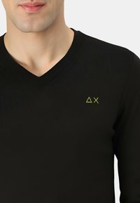 Maglione nero a V con texture a coste, con un piccolo logo verde ΔX sul lato sinistro del petto. Design aderente.