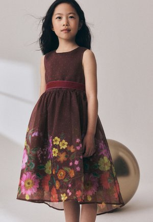 Junges Mädchen mit langen dunklen Haaren, das ein ärmelloses braunes Kleid mit Blumenmuster trägt und vor einem einfarbigen Hintergrund steht.