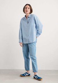 Gai zilgani svītrota krekls ar pogām, brīvā piegriezumā ar kabatu; apvienots ar gaiši ziliem džinsiem un ziliem sporta apaviem ar baltām svītrām un brūnām zālēm.