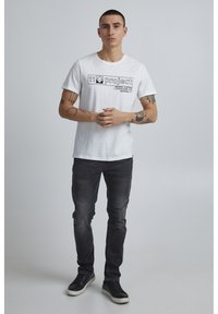 Hvit t-skjorte med korte ermer og svart grafisk tekst, parret med mørkegrå denimjeans og svarte sneakers. Enkelt design med en avslappet passform.