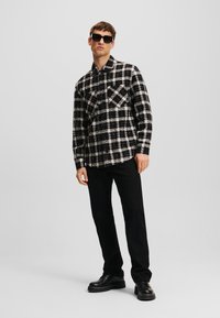 Camisa de cuadros en blanco y negro con botones y bolsillos en el pecho, combinada con pantalones negros y zapatos negros gruesos. Modelo lleva gafas de sol oscuras.