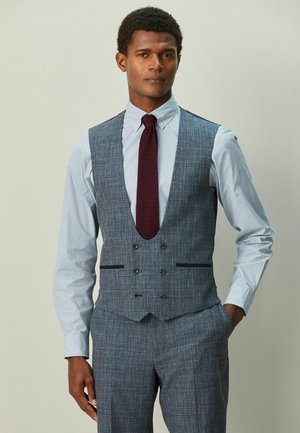 Uomo con camicia azzurro chiaro, gilet grigio a quadri e pantaloni coordinati, indossa una cravatta a maglia rosso scuro, posa con una mano in tasca.