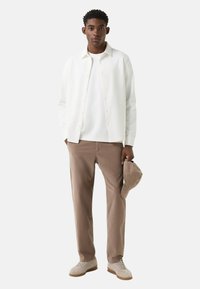 Biała koszula zapinana na guziki nałożona na białą koszulkę, w połączeniu z jasnobrązowymi spodniami. Model trzyma beżową czapkę i nosi jasne sneakersy.