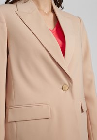 Beige sako s hladkou texturou, zapínání na jeden zlatý knoflík a dvě ploché kapsy. S klopami a otevřeným límcem.