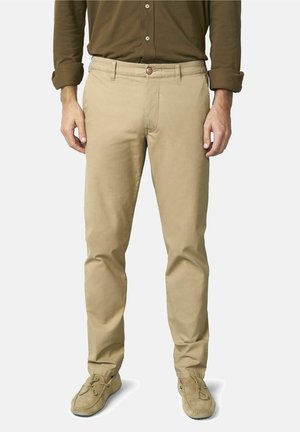 Man draagt beige slim-fit chino's, een bruin overhemd met opgerolde mouwen en beige mocassins, staand tegen een effen achtergrond.