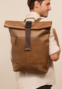 Mochila de cuero marrón con diseño de parte superior enrollable, correas de tela y bolsillo frontal con cremallera. Textura suave y detalles de costura visibles.