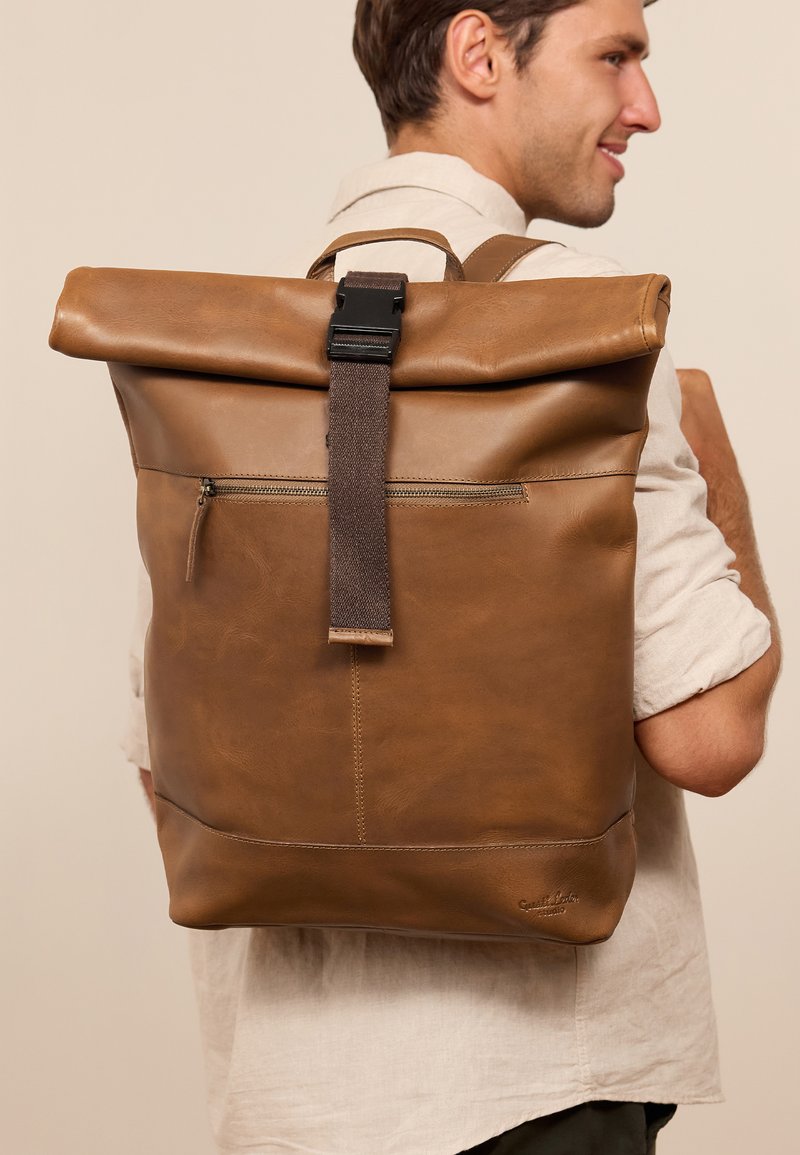 Mochila de cuero marrón con diseño de parte superior enrollable, correas de tela y bolsillo frontal con cremallera. Textura suave y detalles de costura visibles.