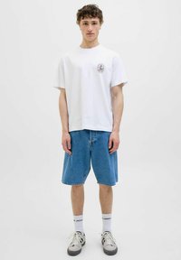 Hvid bomulds t-shirt med et cirkulært grafisk design på venstre bryst, parret med blå denimshorts. Modellen har grå sneakers på med hvide sokker.