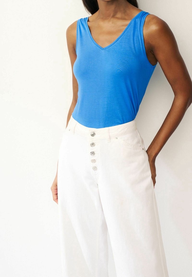 Top blu v-neck senza maniche abbinato a pantaloni bianchi a vita alta con gamba larga, caratterizzati da chiusura con bottone e texture liscia.