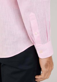 Puño de camisa de lino rosa claro de manga larga, con un diseño puntiagudo, cierre de botón y una textura sutil. Ropa negra visible abajo.