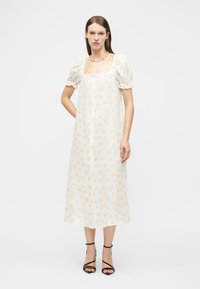 Γυναίκα που στέκεται, φορώντας λευκό μίντι φόρεμα με φουσκωτά μανίκια και μικρό κίτρινο λουλουδάτο σχέδιο, συνδυασμένο με μαύρα πέδιλα με λουράκια και κολιέ.