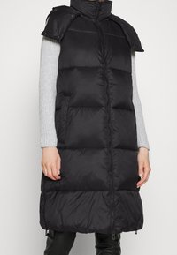 Schwarze Pufferweste mit hohem Kragen, Druckknopf-Schulterakzenten und durchgehendem Reißverschluss. Verfügt über seitliche Taschen und eine gesteppten Textur.