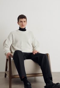 Maglione bianco a coste indossato sopra un dolcevita nero, abbinato a pantaloni neri. La persona è seduta su una sedia in legno minimalista.