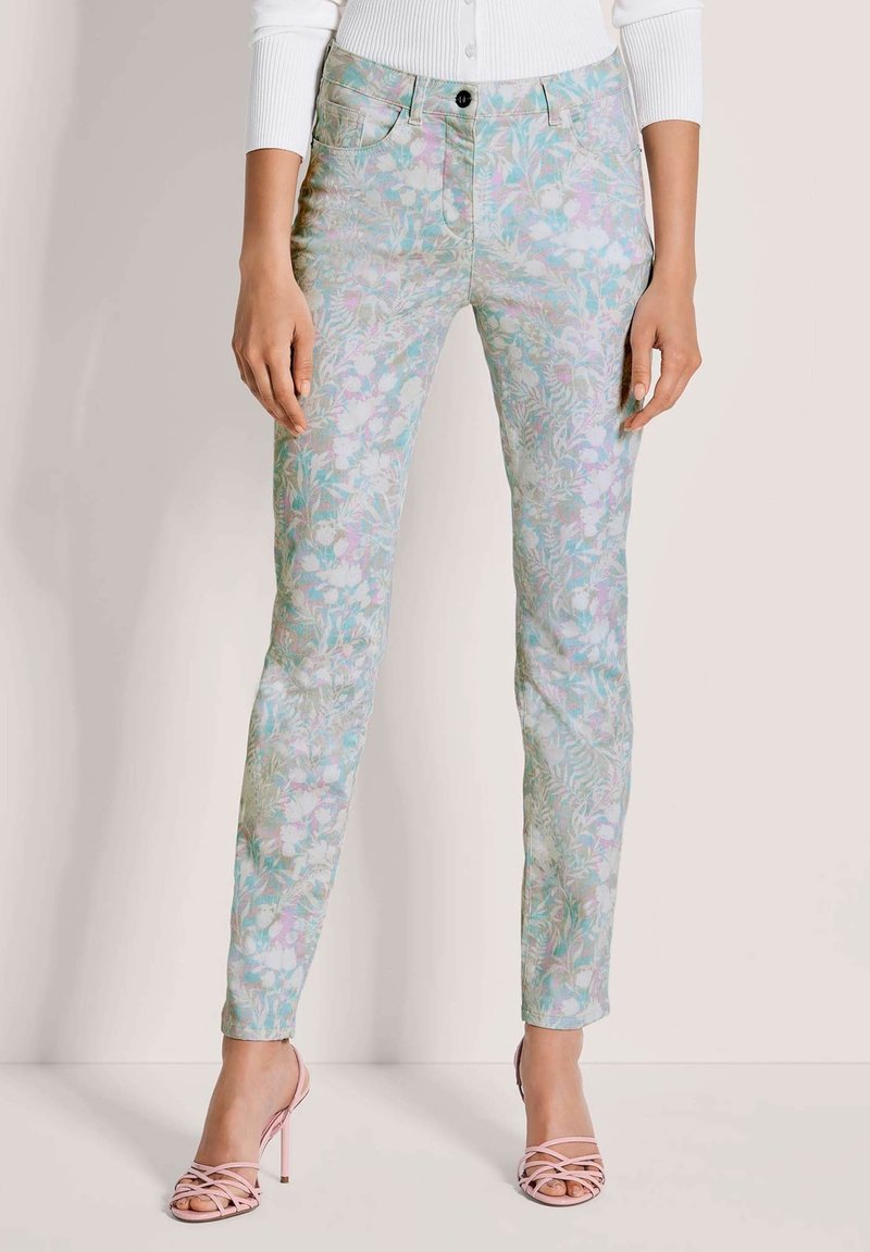 Pantalon slim à motifs floraux pastel porté avec un haut côtelé blanc et des sandales à talons hauts roses à lanières, debout devant un fond uni clair.