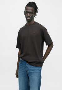 Fiatal férfi dreadlock stílusú hajjal, sötét, túlméretezett pólót és kék farmert viselve, egyik kezét a zsebében tartva, egyszínű háttér előtt áll.