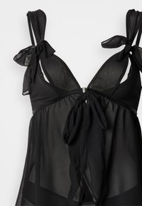 Haut de lingerie noire transparente avec bretelles réglables en forme de nœud, décolleté plongeant et détail de taille nouée. Tissu léger au toucher lisse.