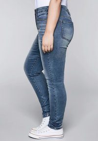 Úzké modré džíny z vybledlého denimu s klasickým pětikapsovým designem. Kombinovány s bílými vysokými teniskami.