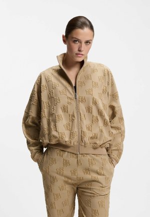 Mujer con las manos en los bolsillos, vistiendo una chaqueta beige con cremallera y pantalones a juego, ambos con un patrón repetido y texturizado del logo en relieve.