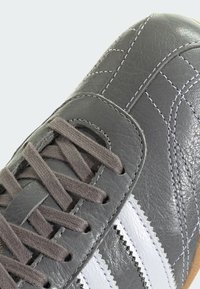 Γκρι δερμάτινο αθλητικό παπούτσι με λευκές ρίγες, επίπεδα γκρι κορδόνια, υφή στην επιφάνεια και ρα�φές. Φαίνεται η λαστιχένια σόλα στο κάτω μέρος.