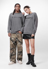 Felpe grigie oversize con dettagli del logo neri, pantaloni a motivo mimetico e una mini gonna di pelle nera. La calzatura include sneakers bianche e stivali neri.