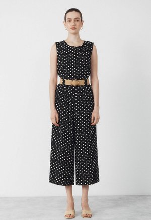 Kobieta w czarnym kombinezonie bez rękawów w białe kropki, z szerokim beżowym paskiem, złotymi kolczykami kołami i beżowymi butami na obcasie z odkrytymi palcami.