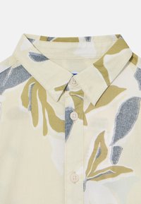 Camisa de manga corta con un fondo beige claro y grandes patrones florales en verde y gris, con cuello tipo camisa y cierre de botones.
