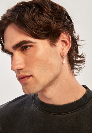 Boucle d'oreille en argent avec une perle pendante. Le sujet a des cheveux châtains ondulés de longueur moyenne et porte un sweat-shirt gris foncé à encolure arrondie.
