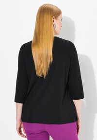 Zwarte top met driekwart mouwen van een gladde stof, gecombineerd met paarse broek, van achteren weergegeven tegen een witte achtergrond.