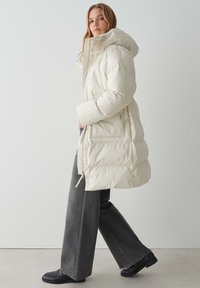 Doudoune blanche cassée avec capuche, dotée d'une fermeture éclair à l'avant, de poches latérales et d'un design matelassé. Associée à un jean gris à larges jambes et à des chaussures noires.