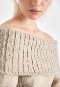 Beige off-shoulder tröja med ribbat mönster, som har en vid halsringning och subtila horisontella ränder i ett mjukt stickat material.