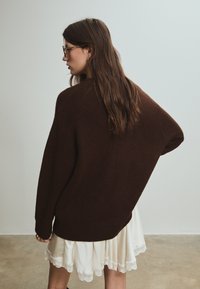 Maglione marrone oversize con una texture morbida, scollo rotondo e spalle scese, abbinato a una gonna bianca con bordo in pizzo.