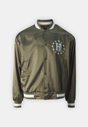 Olivengrønn sateng bomberjakke med en sirkulær logo med stjerner og bokstaven "H", trykknapper, samt stripete ribbestrikkede mansjetter og krage.