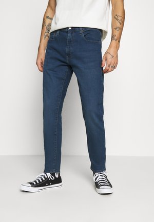 Schmale blaue Denim-Jeans mit einem leichten Stretch. Verfügt über ein klassisches Fünf-Taschen-Design und einen Metallknopfverschluss. Getragen mit schwarzen Turnschuhen.