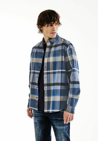 Camicia a quadri blu con bottoni, caratterizzata da diverse tonalità di blu, beige e nero, realizzata in un tessuto leggero. Indossata con una maglietta scura e jeans.