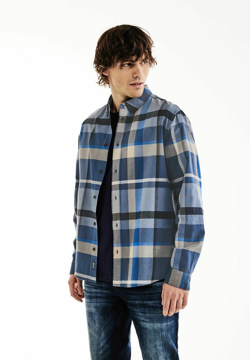 Camicia a quadri blu con bottoni, caratterizzata da diverse tonalità di blu, beige e nero, realizzata in un tessuto leggero. Indossata con una maglietta scura e jeans.