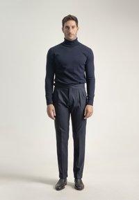 Hombre de pie, de frente, vistiendo un suéter de cuello alto azul marino, pantalones azul marino con plisados y zapatos de cuero negros, sobre un fondo sencillo.