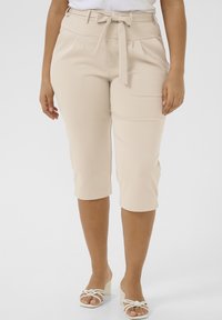 Pantalons beiges, coupés à mi-mollet, avec une ceinture à nouer et des plis à la taille, fabriqués dans un tissu lisse et extensible. Portés avec des sandales blanches.