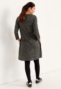 Robe à motifs noir et doré avec des manches trois-quarts, longueur genou, poches latérales, associée à des leggings noirs et des chaussures de sport.