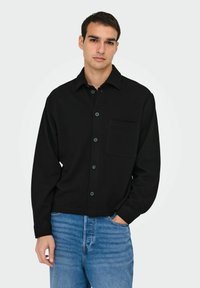 Black button-up shirt with a collar, single chest pocket, and long sleeves. Worn with light blue jeans, displayed against a neutral background.