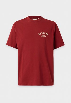 Un mannequin masculin porte une casquette noire, un t-shirt rouge avec un motif graphique, un jean bleu, une ceinture marron et des baskets pâles sur un fond blanc en studio.