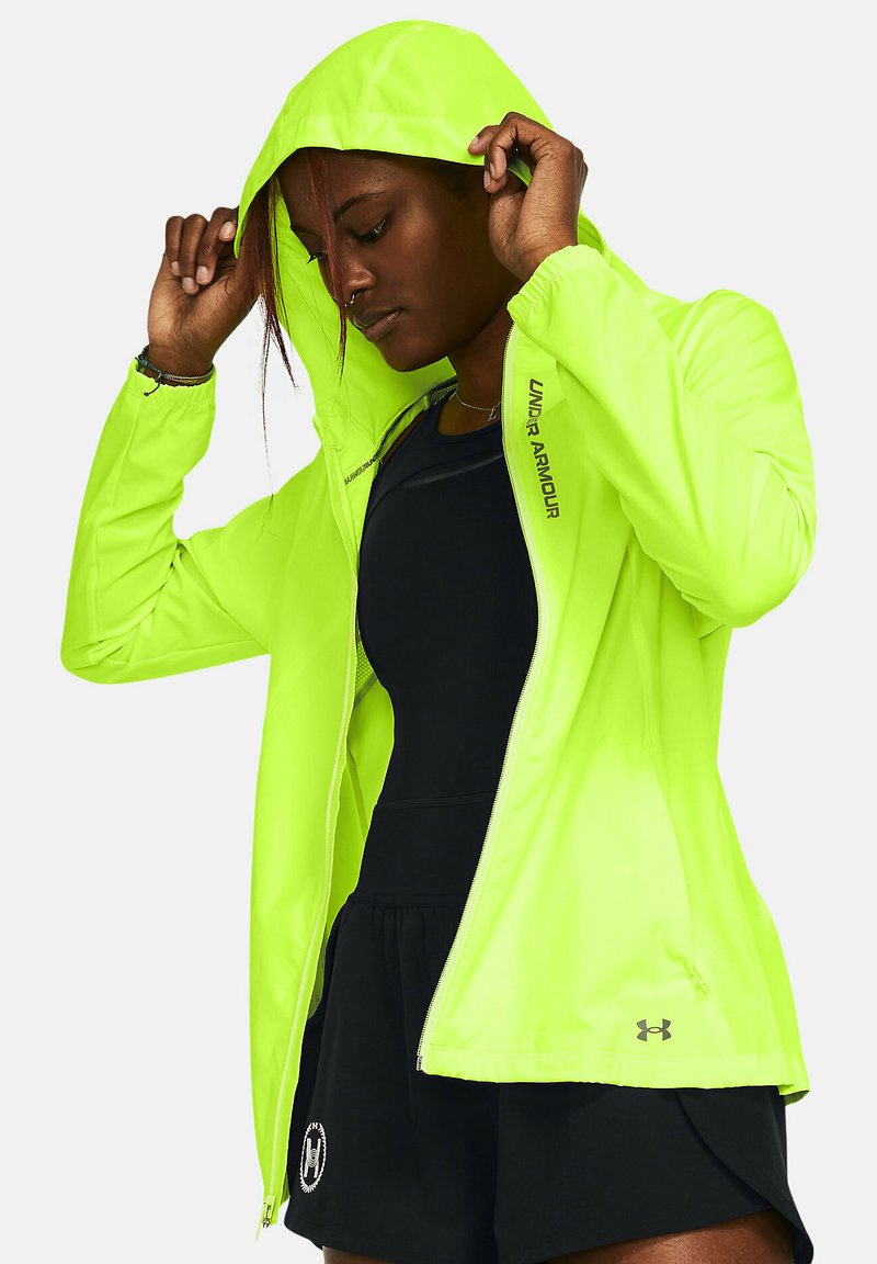 Giacca leggera con cappuccio di colore verde neon, con cerniera, maniche lunghe e logo Under Armour. Indossata sopra un top sportivo nero e pantaloni corti.