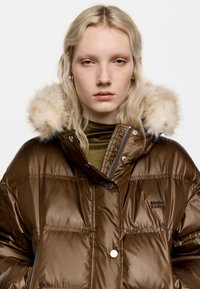 Hnedý páperový kabát s lesklým povrchom, ktorý má kapucňu z umelej kožušiny, zips a patentky, a prešívanú textúru. Logo na hrudi.