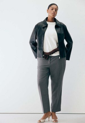Femme portant une veste en cuir noire, un haut blanc, un pantalon à carreaux gris et des talons ouverts beiges sur un fond blanc uni.