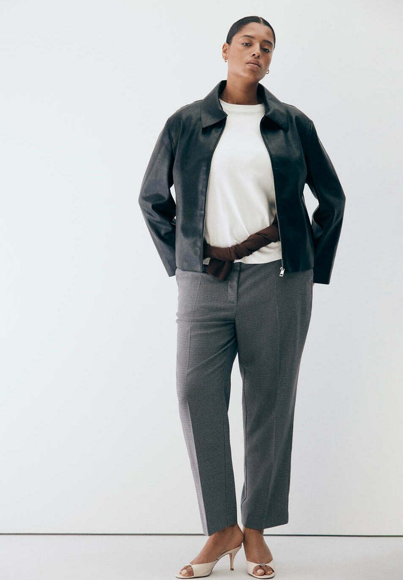 Femme portant une veste en cuir noire, un haut blanc, un pantalon à carreaux gris et des talons ouverts beiges sur un fond blanc uni.