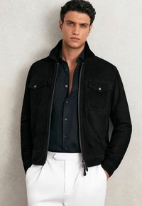 Melnas zamša jakas ar rāvējslēdzēja aizdari un divām krūšu kabatām; kopā ar tumši zilu krekli un baltām biksēm, fona fonā neitrāla krāsa.